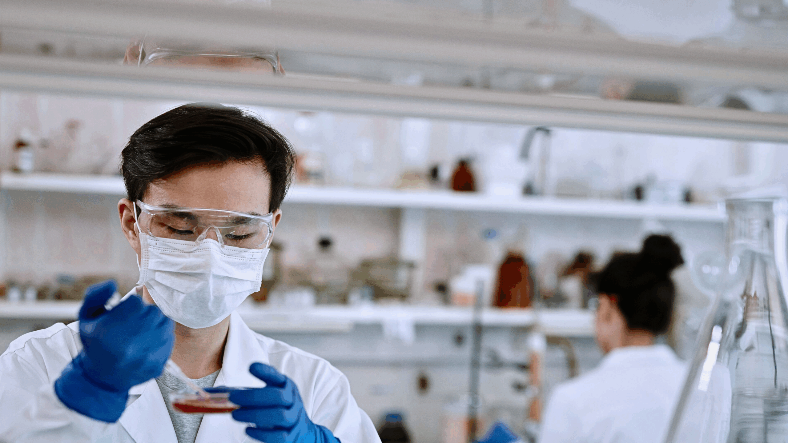 Researcher in lab coat with eye protection, gloves, and facemask, pipettes into a petri dish in a laboratory with a second researcher in the background.